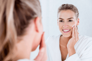 Smiling woman applying moisturizer on her face in front of a mirror