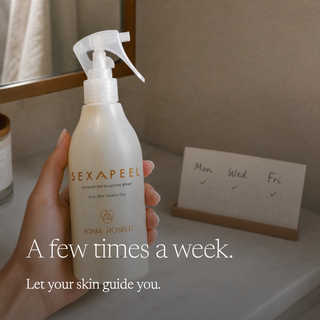 A hand holds a bottle of SEXAPEEL Instant Revolution Spray by Sona Roselli in a bathroom setting. The bottle is off-white with gold lettering, and the background shows a tiled wall, a mirror, and a small weekly planner card on a marble counter.