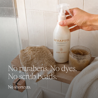 A hand holds a bottle of Nona Roselli Sexapeel Exfoliating Spray in a bathroom setting. The scene includes a wooden shelf with a beige scrubber, a jar of scrub, and a white towel, with text overlay reading 'No parabens.