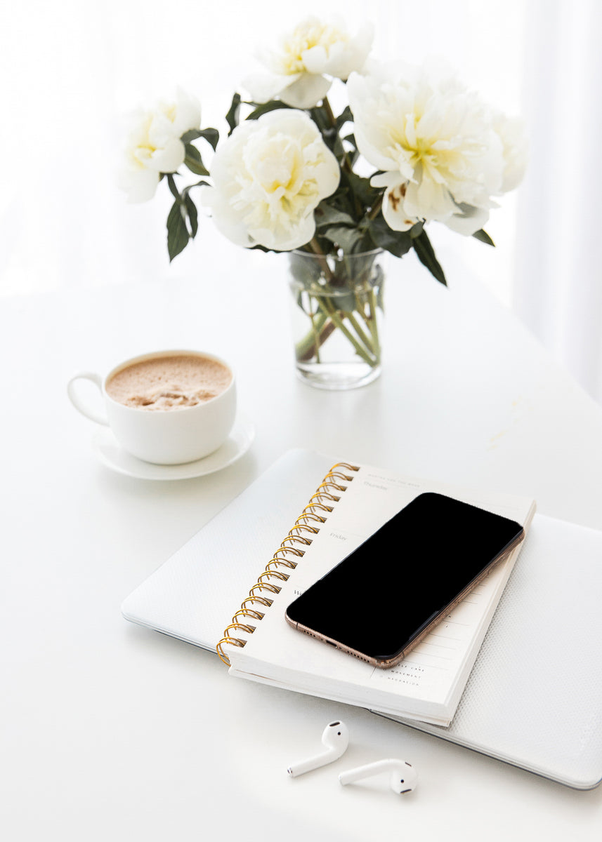 A phone on a notebook. They are resting on a macbook next to airpods. A cup of coffee to the left and a bouquet of white beautiful flower.