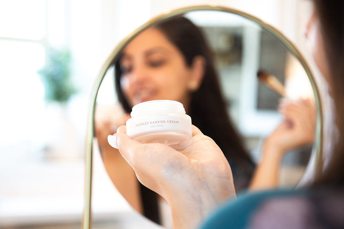 Woman looking down at Intense Barrier Cream moisturizer in front of a mirror.