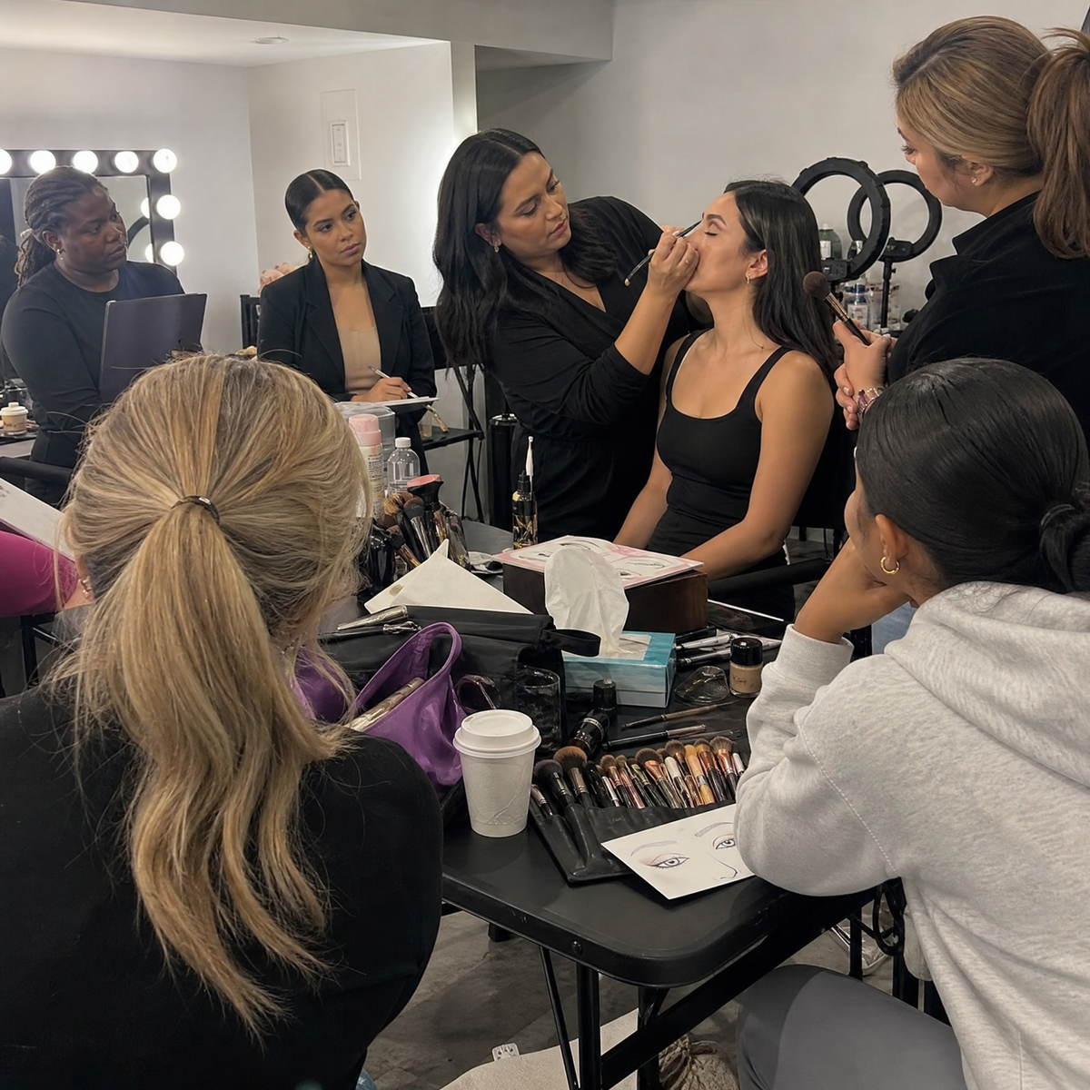 A makeup artist applies skin prep and makeup to a seated model in a bright makeup room, with other stylists and makeup tools visible around the table.