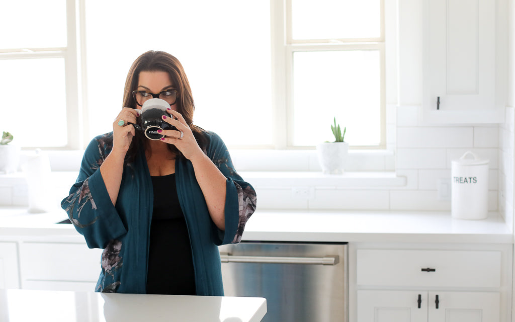 Sonia Roselli stands in her cozy kitchen, savoring a moment as she takes a sip from a coffee cup, exuding warmth and relaxation in her home environment.
