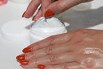 Close-up of hands with glossy skin and polished nails opening a jar of rich white cream, showing the product’s smooth, hydrating texture.