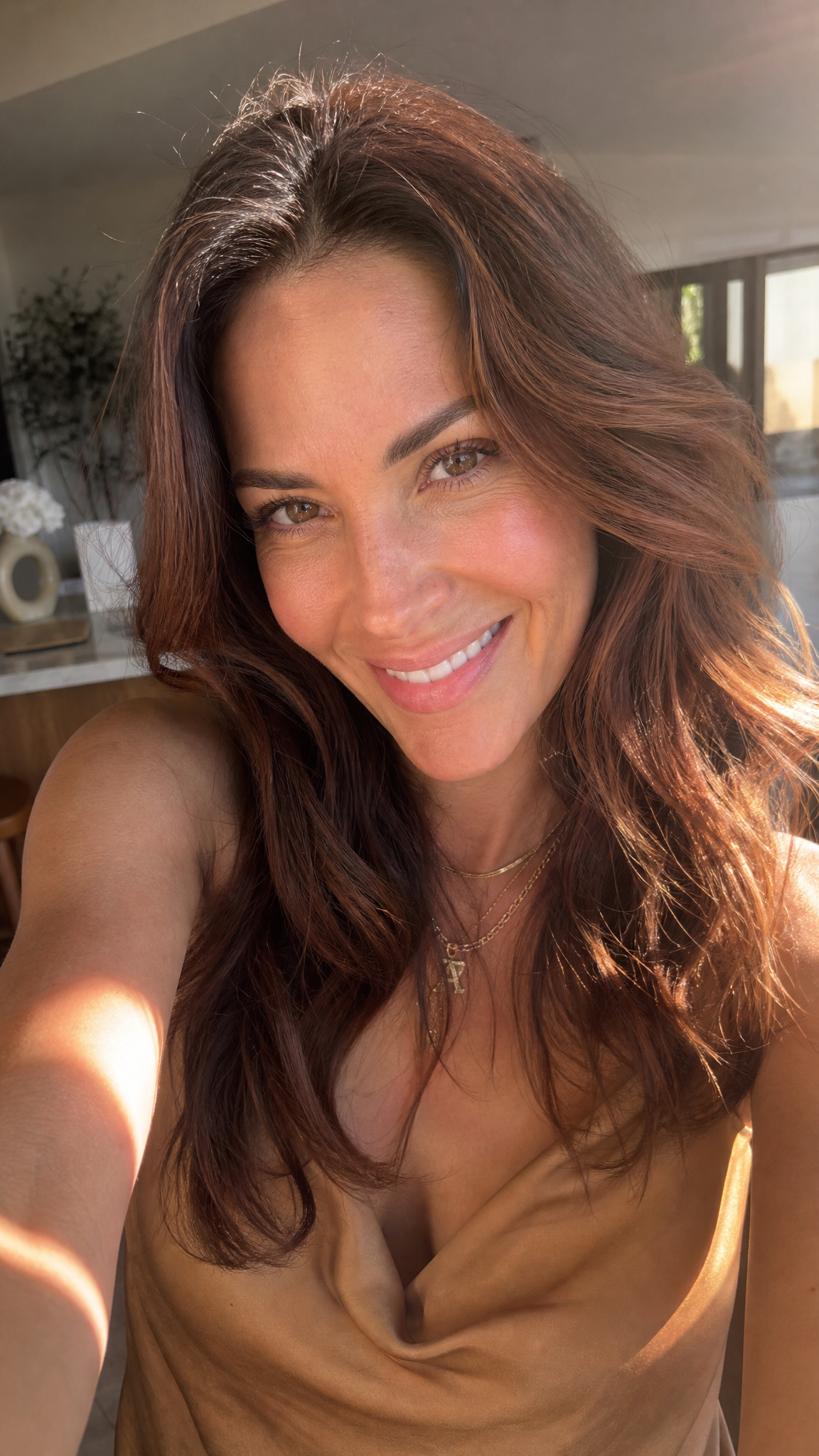 A smiling woman with long brown hair wearing a brown draped top, taking a selfie in a brightly lit indoor setting.