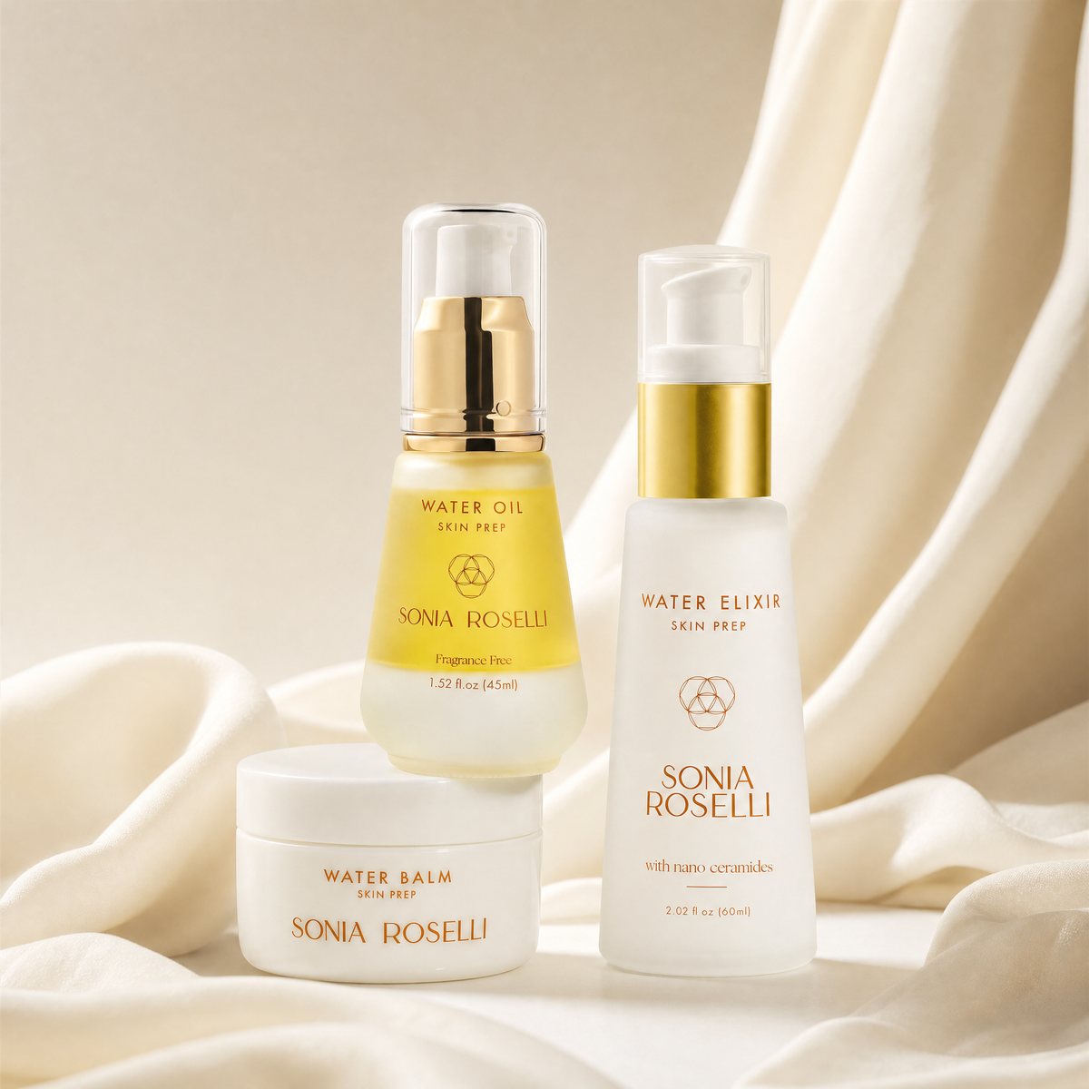 Three Sonia Roselli skincare products arranged on draped cream fabric: a Water Oil pump bottle with a gold cap, a Water Balm jar with a white lid, and a Water Elixir pump bottle with a gold cap.