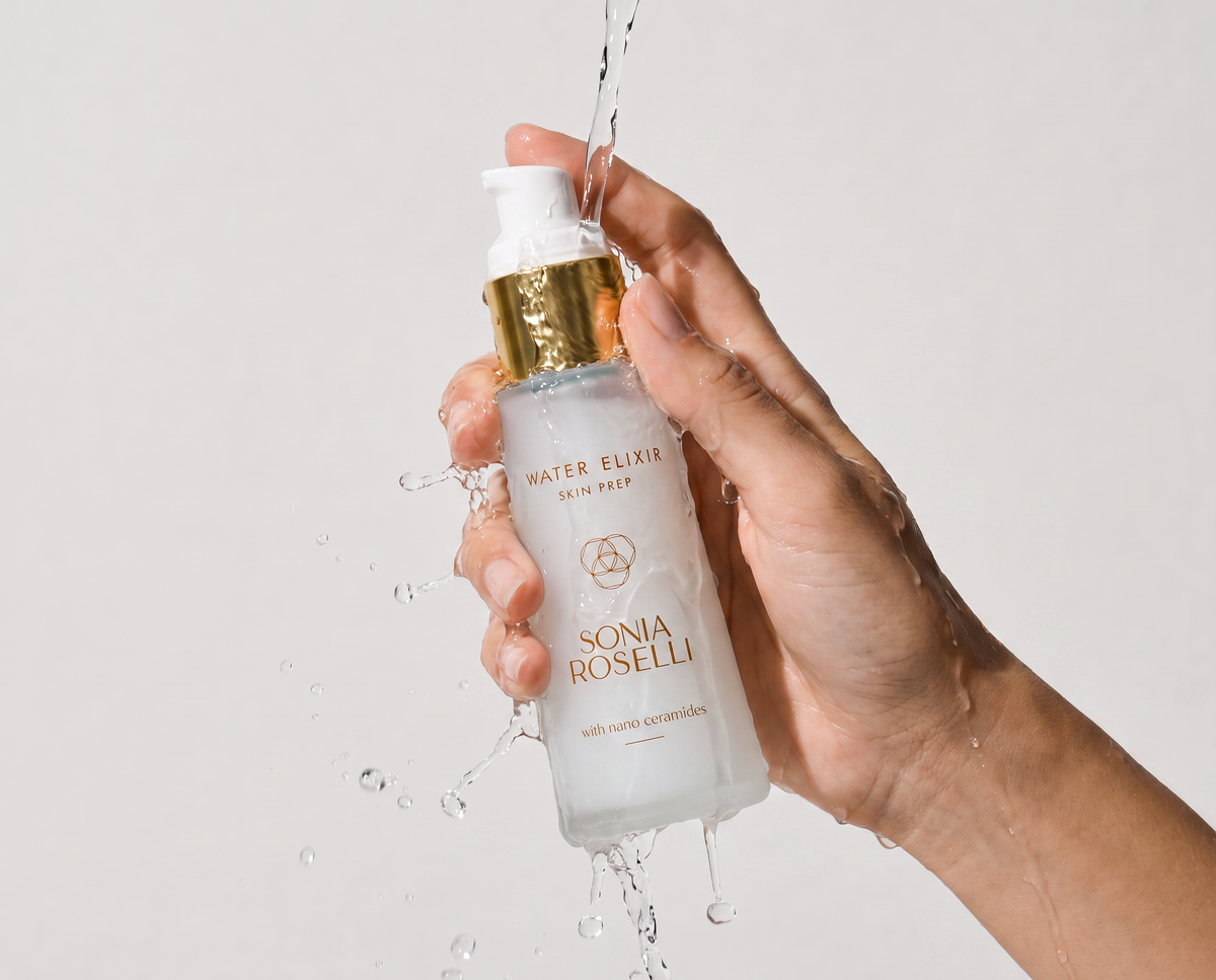 A hand holding a frosted glass pump bottle of Sonia Roselli Water Elixer Sun Serum, with water splashing around it against a light gray background.