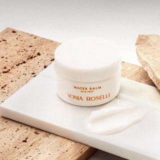 A small white jar labeled 'Water Balm Skin Prep' by Sonia Roselli, placed on a white surface with a quantity of the product spread out in front of it, against a backdrop of textured stones. Nourishing balm that seals in hydration and protects skin barrier - lightweight moisturizer for dry, sensitive skin Gel cream moisturizer for skin that feels tight and dry - lightweight hydration that actually lasts