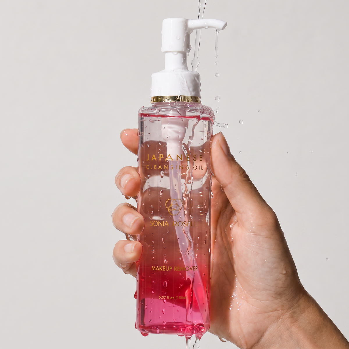 A hand holding a clear pump bottle of Sonia Roseletti Japanese Cleanse Oil makeup remover, with water dripping from the pump and droplets on the bottle, against a light gray background.