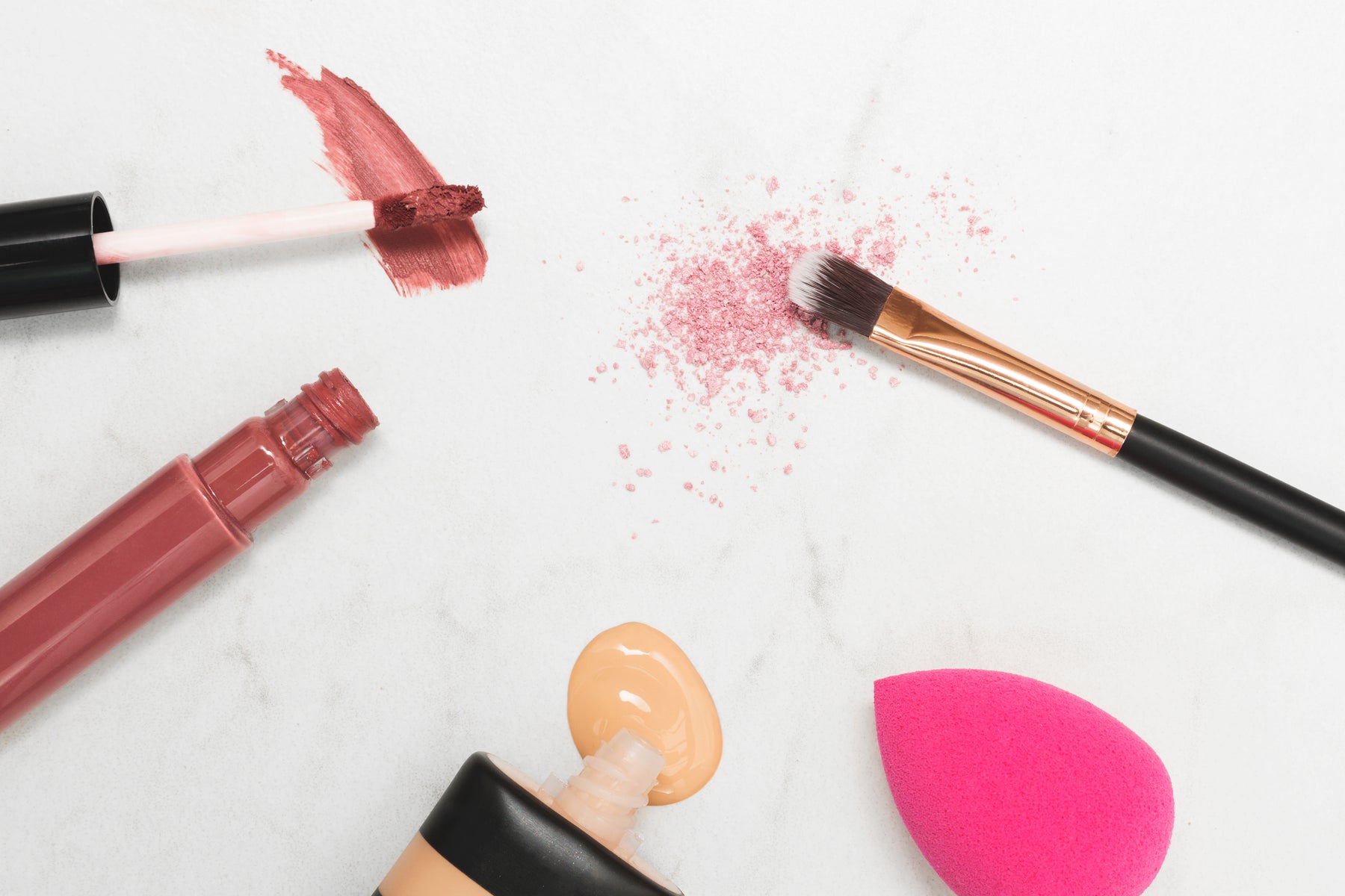 Makeup brush, a makeup sponge, foundation, and a lipstick on marble background