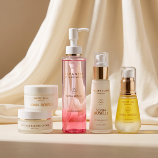 Five Sonia Roselli skincare bottles and jars arranged together on a draped cream fabric backdrop. The set includes a pink pump bottle, a white pump bottle, a yellow oil bottle, and two white cream jars.