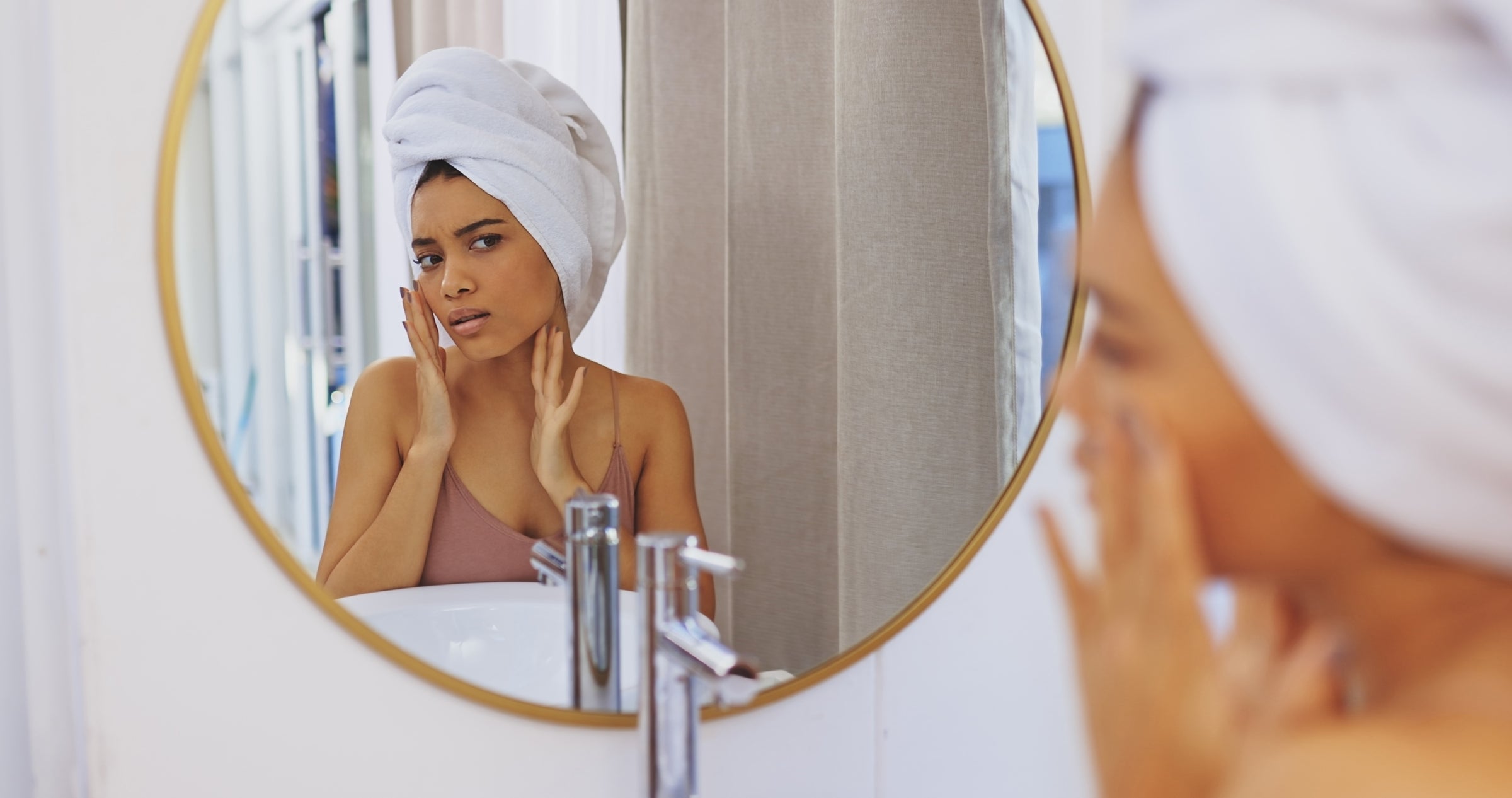 Woman looking in mirror with a towel on her head. Her hands are on her face.