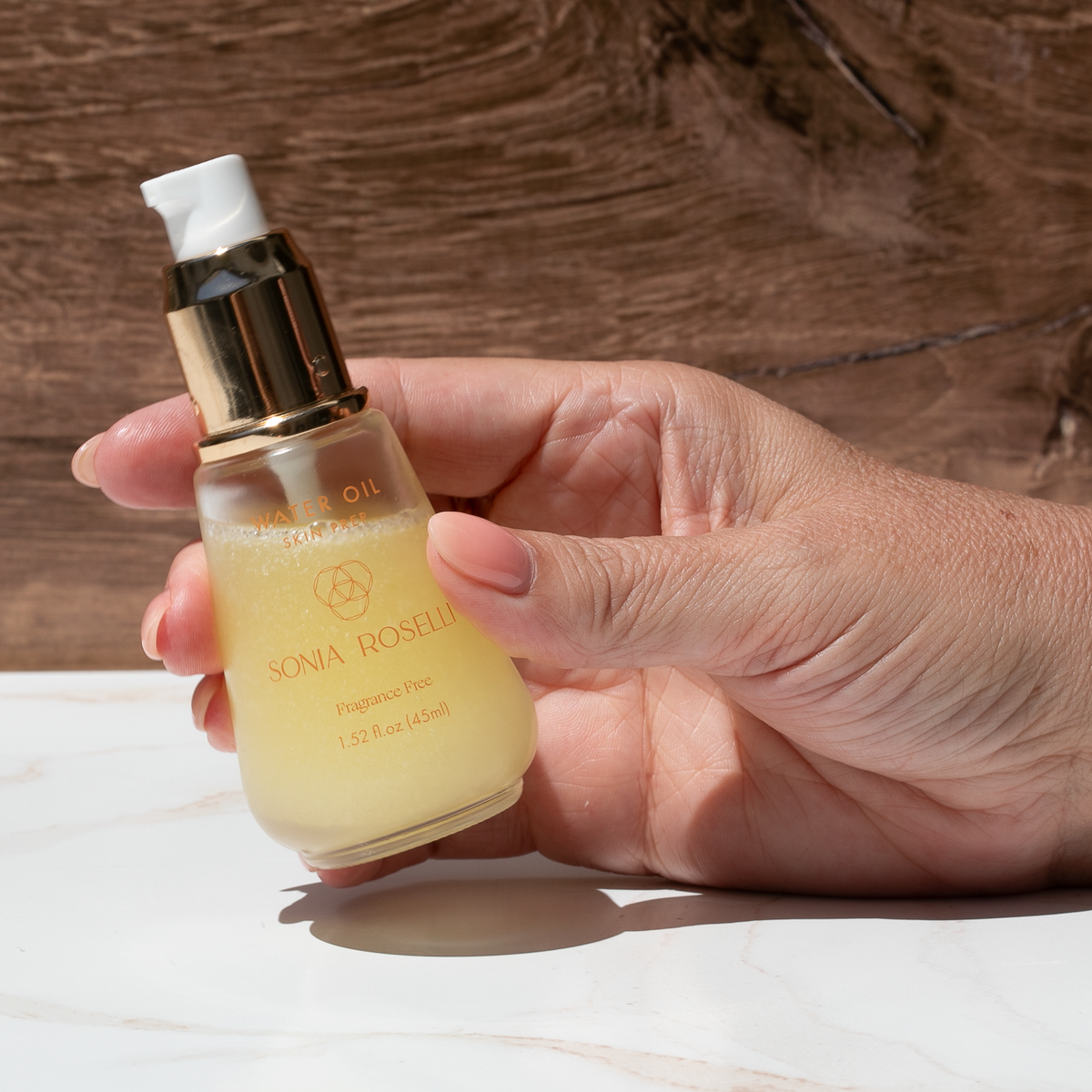 A hand holding a small glass bottle of Sonia Roselli Fragrance Oil with a gold pump and white cap, set against a rustic wooden background.