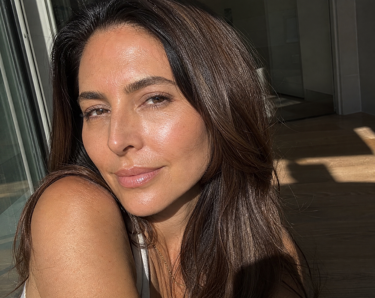 A close-up portrait of a woman with long dark brown hair, wearing a white top, set against a modern interior background with warm sunlight and shadows.