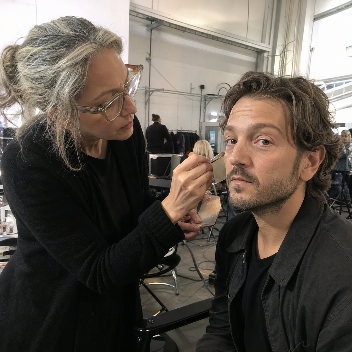 A makeup artist with gray hair and glasses applies makeup to a man's face in a backstage studio setting.