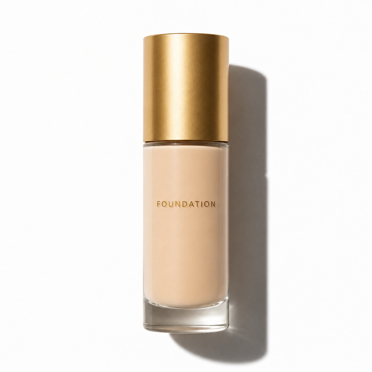 A clear glass bottle of liquid foundation with a metallic gold cap, filled with a light beige foundation, and labeled 'FOUNDATION' in gold lettering, set against a clean white background with a soft shadow.