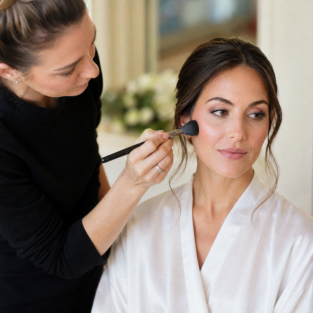 A bridal makeup artist applies makeup to a woman's face who is a bride using a brush, with the woman wearing a white satin robe in a softly lit indoor setting.