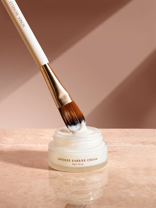 Skincare brush dipped in jar of intense barrier cream with marble cunter and tan background