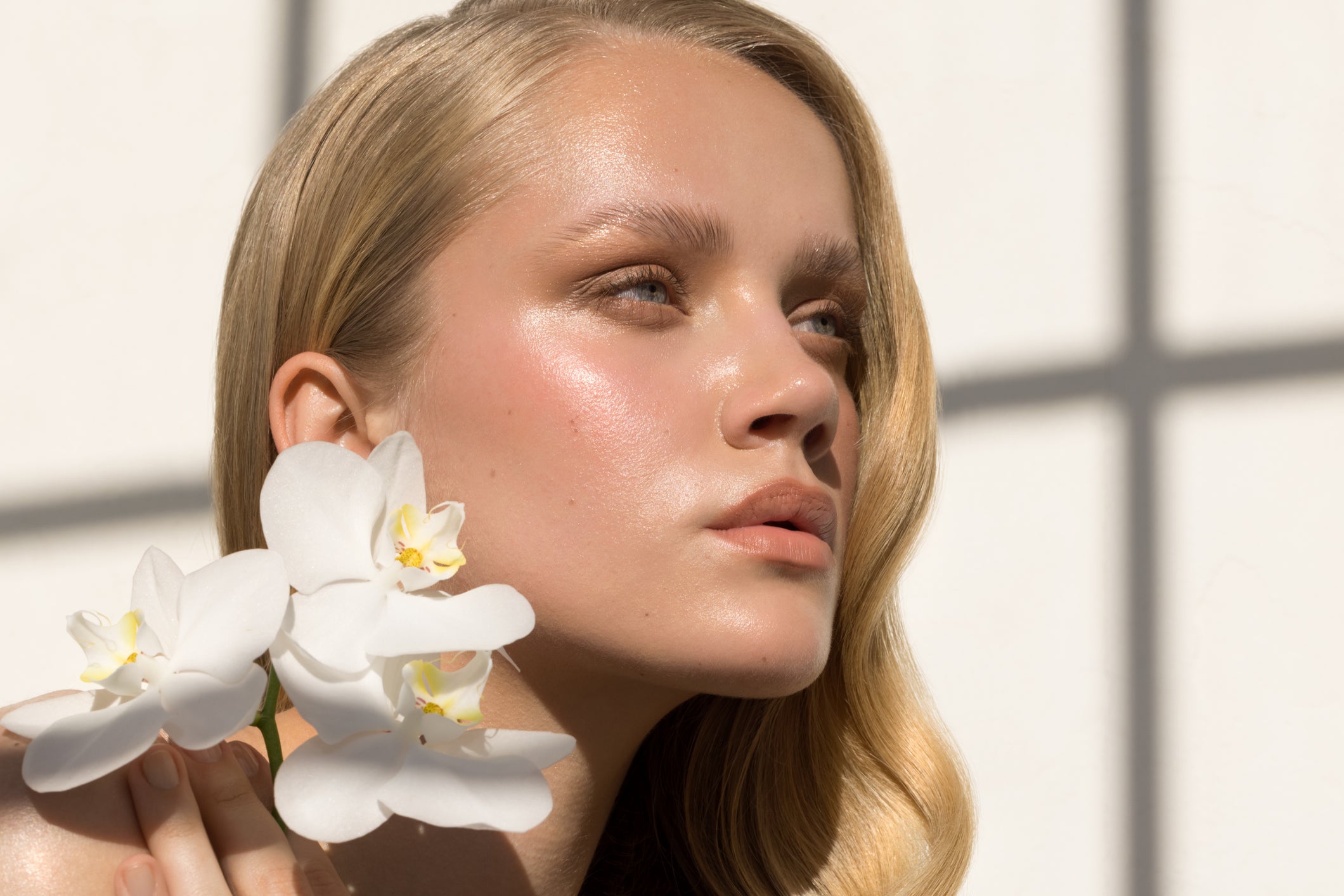 A women with glowing skin, holding orchids near her shoulder, looks into the distance 