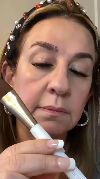 Woman holding a skincare brush near her face with eyes closed, wearing a jeweled headband and hoop earrings, demonstrating skin prep technique