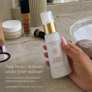 A hand holds a bottle of Sonia Roselli Water Elixir Skin Prep against a beige marble countertop with makeup items in the background.
