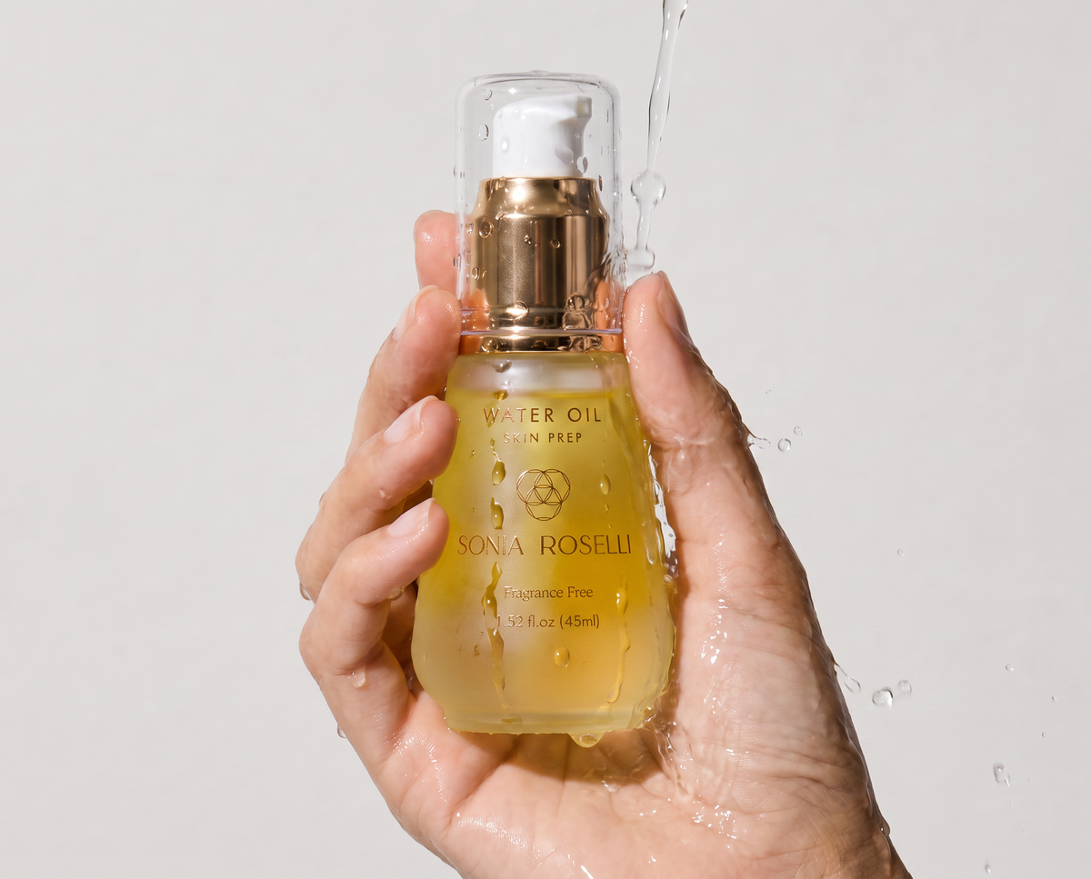 A hand holding a bottle of Sona Roselli Water Oil Skin Prep serum, with water droplets and a stream of water splashing around it against a light gray background.