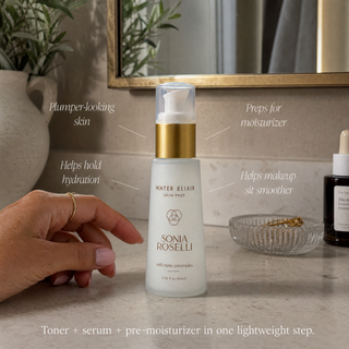 A hand holds a frosted white pump bottle of Sonia Roselli Water Elixir Skin Prep on a bathroom counter, with text callouts describing its benefits. A mirror, plants, and a glass dish are visible in the background.